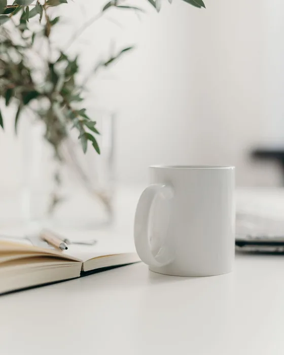 A plain white mug on an office desk beside a plant and an open notebook.