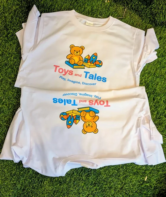 White T-shirts printed with the colourful Toys and Tales logo, laid on grass.