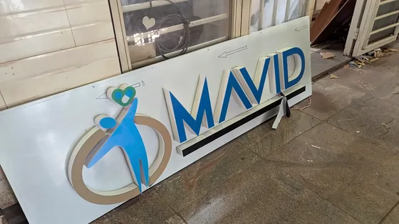White sign panel with a cut-out MAVID logo in blue and gold, leaning against a window.