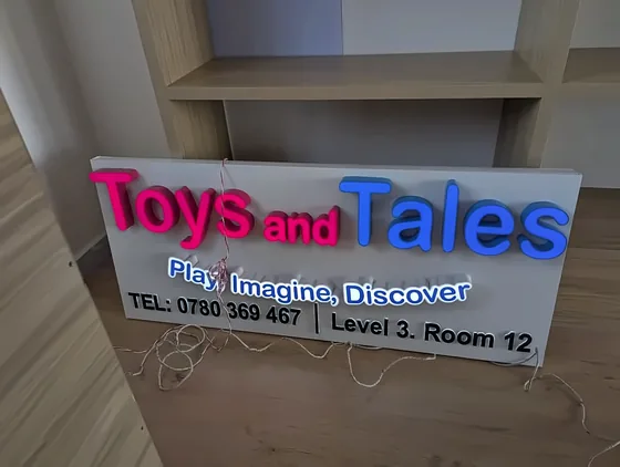 Illuminated sign with pink and blue Toys and Tales lettering above a printed contact panel.